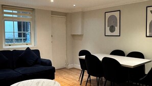 Una sala de estar moderna con un sofá azul oscuro, una mesa de centro redonda, una mesa de comedor con sillas negras y una ventana con persianas.