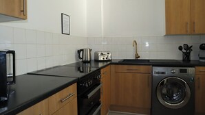 A modern kitchen with a black countertop, stainless steel appliances, wooden cabinets, and a built-in washing machine. - BnBNook - Soho and Regent’s Park - 1BR with Balcony (Greater London)