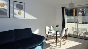 A modern living room with a dark blue sofa, a glass dining table with white chairs, and two framed abstract artworks on the wall. - BnBNook - Soho and Regent’s Park - 1BR with Balcony (Greater London)