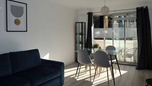 A modern living room with a dark blue sofa, a glass dining table with four chairs, and a large window with a view of a balcony. - BnBNook - Soho and Regent’s Park - 1BR with Balcony (Greater London)