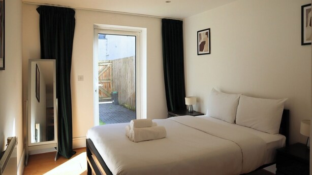A bedroom with a bed, a nightstand, a lamp, a window with curtains, and a view of the outside. - BnBNook - Camden 2BR with Terrace - King's Cross (Greater London)