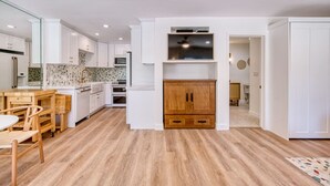 Cozinha moderna com armários brancos, mesa de jantar de madeira e televisão embutida.