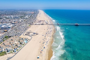 A coastal cityscape with a beach, a pier, and a densely built urban area with buildings and parked cars.