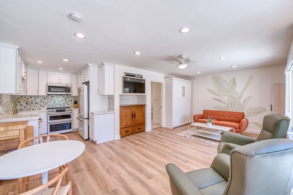 A modern apartment with a kitchen, dining area, and living room. It features white cabinetry, a wooden dining table, and a sofa.