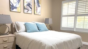 A bedroom with a bed, two bedside tables, a window with blinds, and abstract art on the wall.