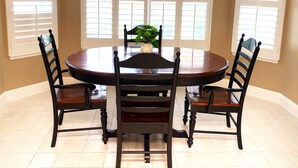 A dining room with a round table, four chairs, a chandelier, and large windows with blinds.
