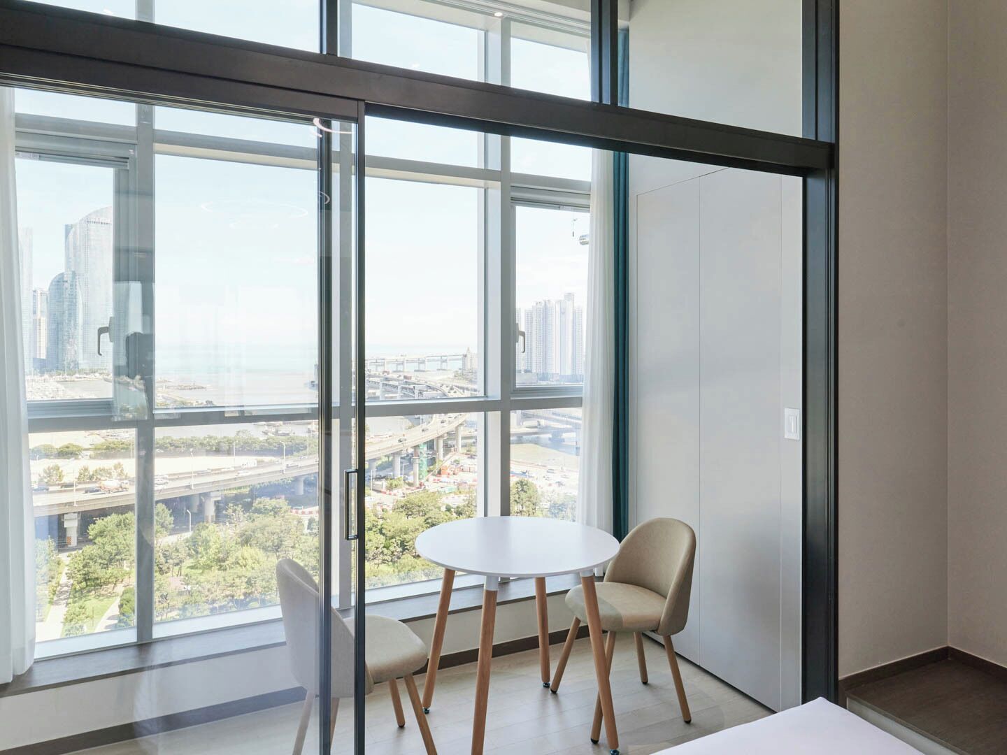 A modern room with large floor-to-ceiling windows, a round table, two chairs, and a view of a cityscape with buildings and a bridge.