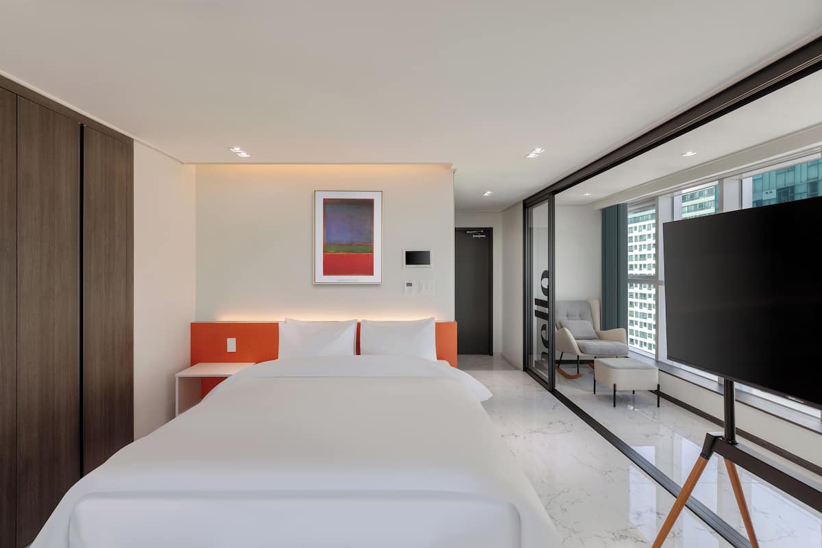 A modern hotel room with a large bed, a flat-screen TV, a seating area with a chair and ottoman, and a large window offering a city view.