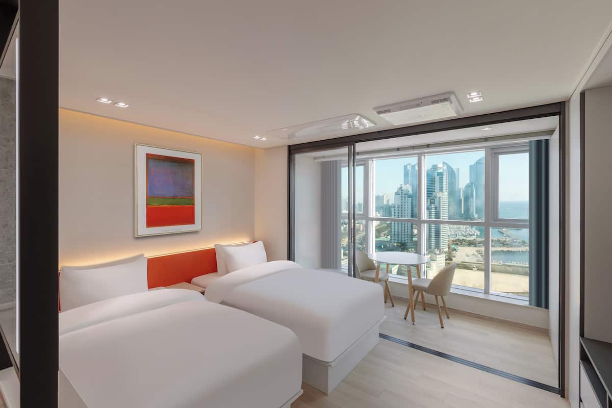 A modern hotel room with two beds, a city view, and a small dining area.
