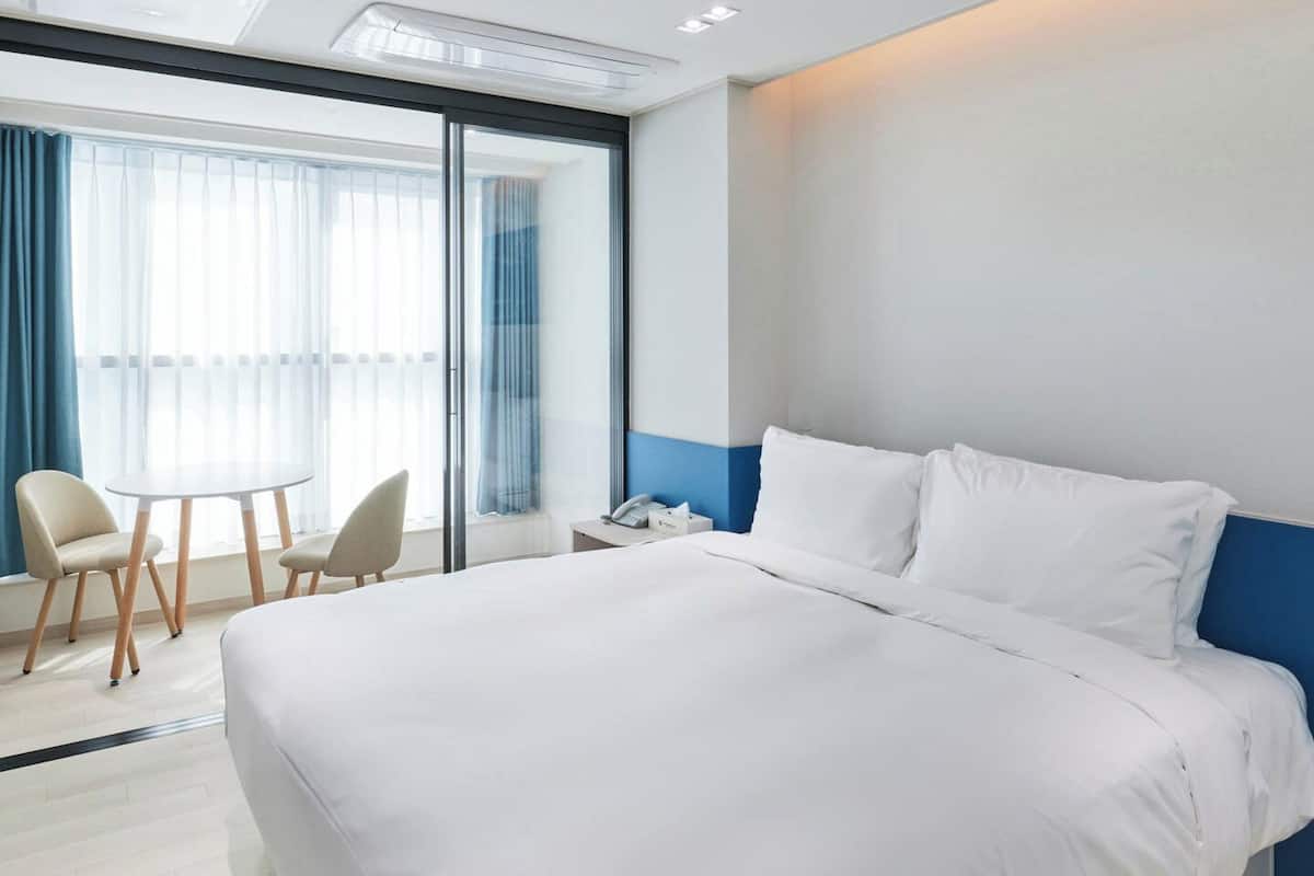 A hotel room with a large bed, a desk, two chairs, and a view of the outdoors through a large window.