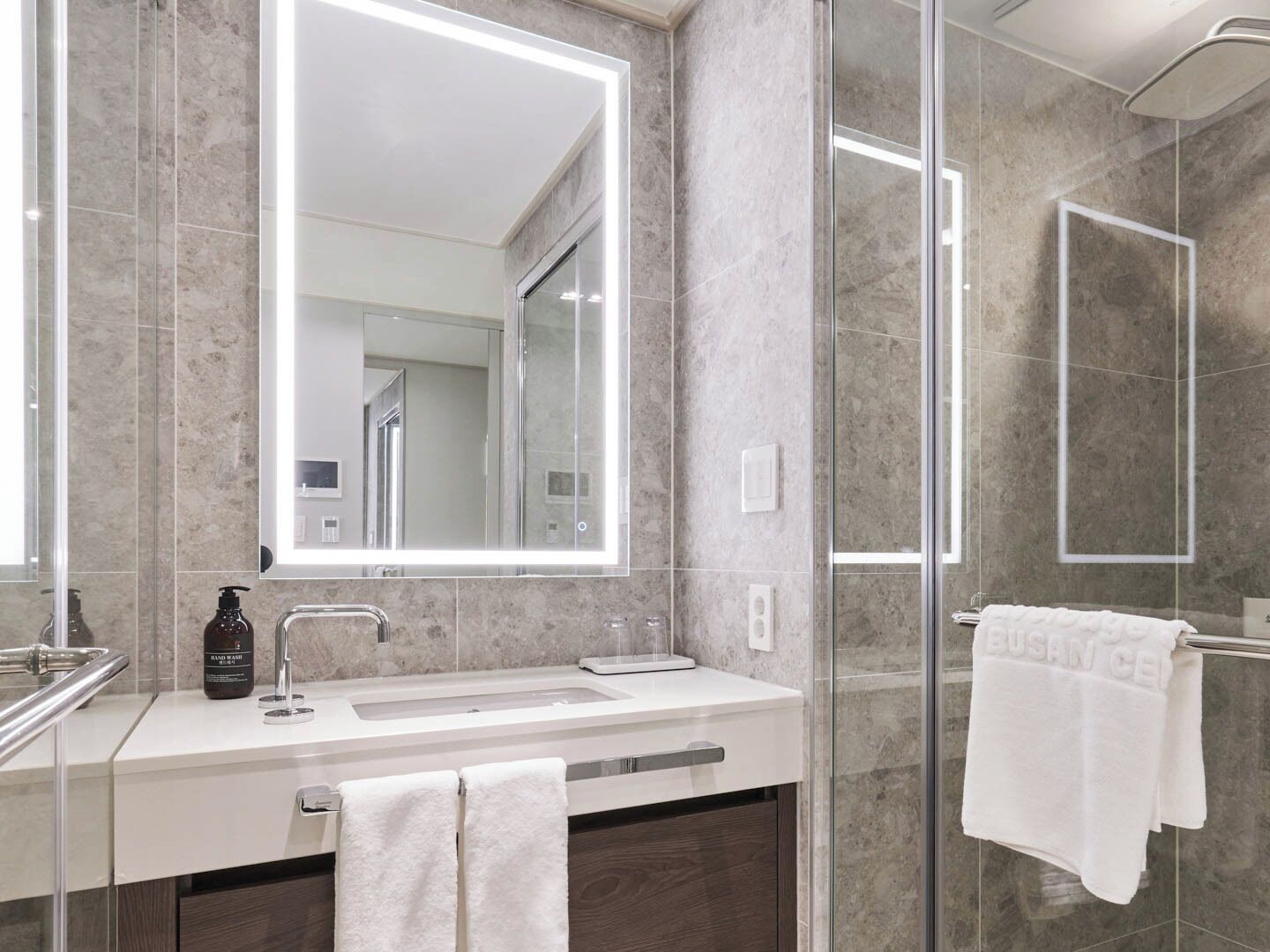 Une salle de bain moderne avec une douche à l’italienne en verre, un grand miroir et un plan de toilette blanc.