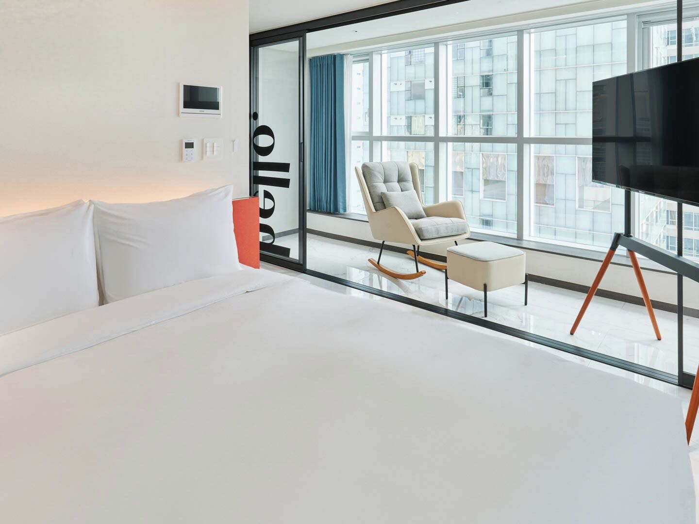 A modern hotel room with a large bed, a seating area with a chair and ottoman, a flat-screen TV, and a large window with a city view.