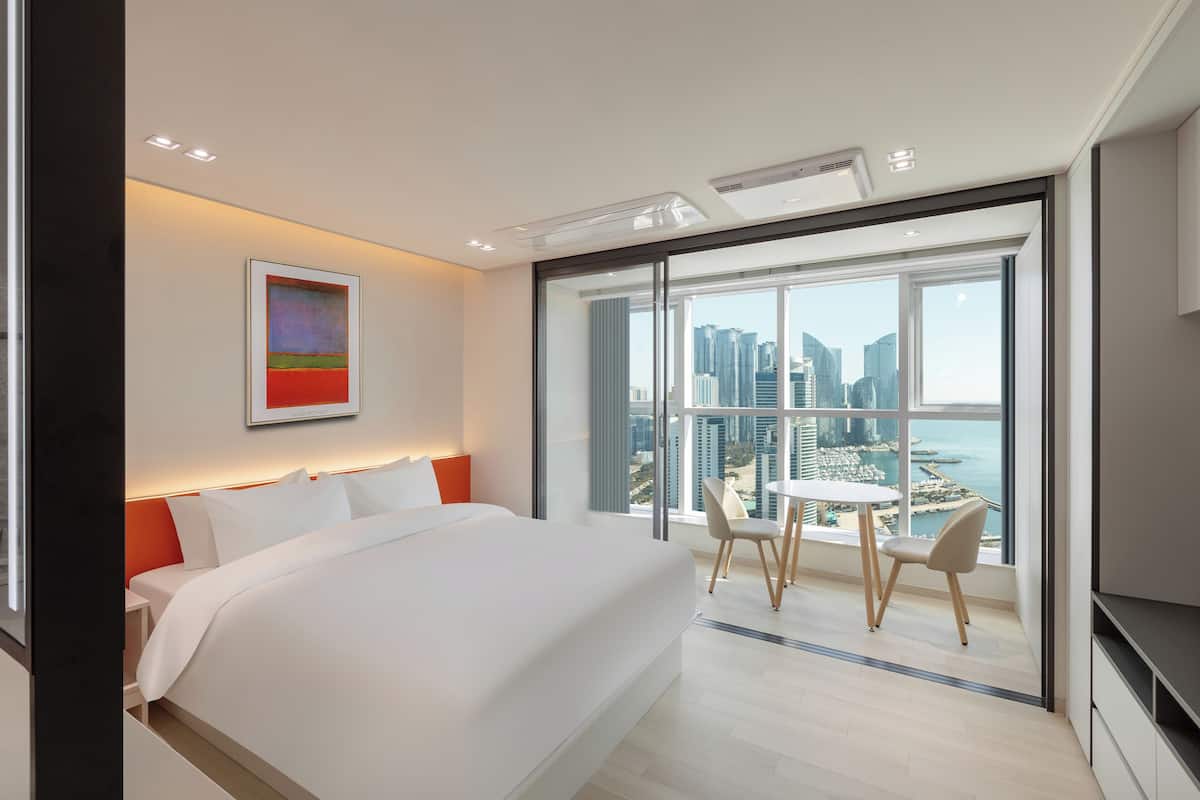 A modern hotel room with a large bed, a dining table, and a city view through large windows.