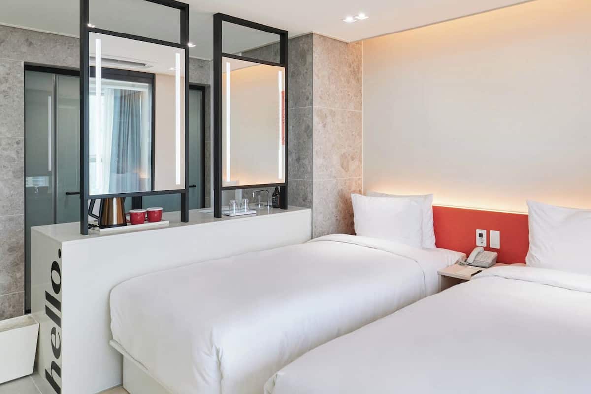 A modern hotel room with two single beds, a white vanity with a mirror, and a red accent wall.