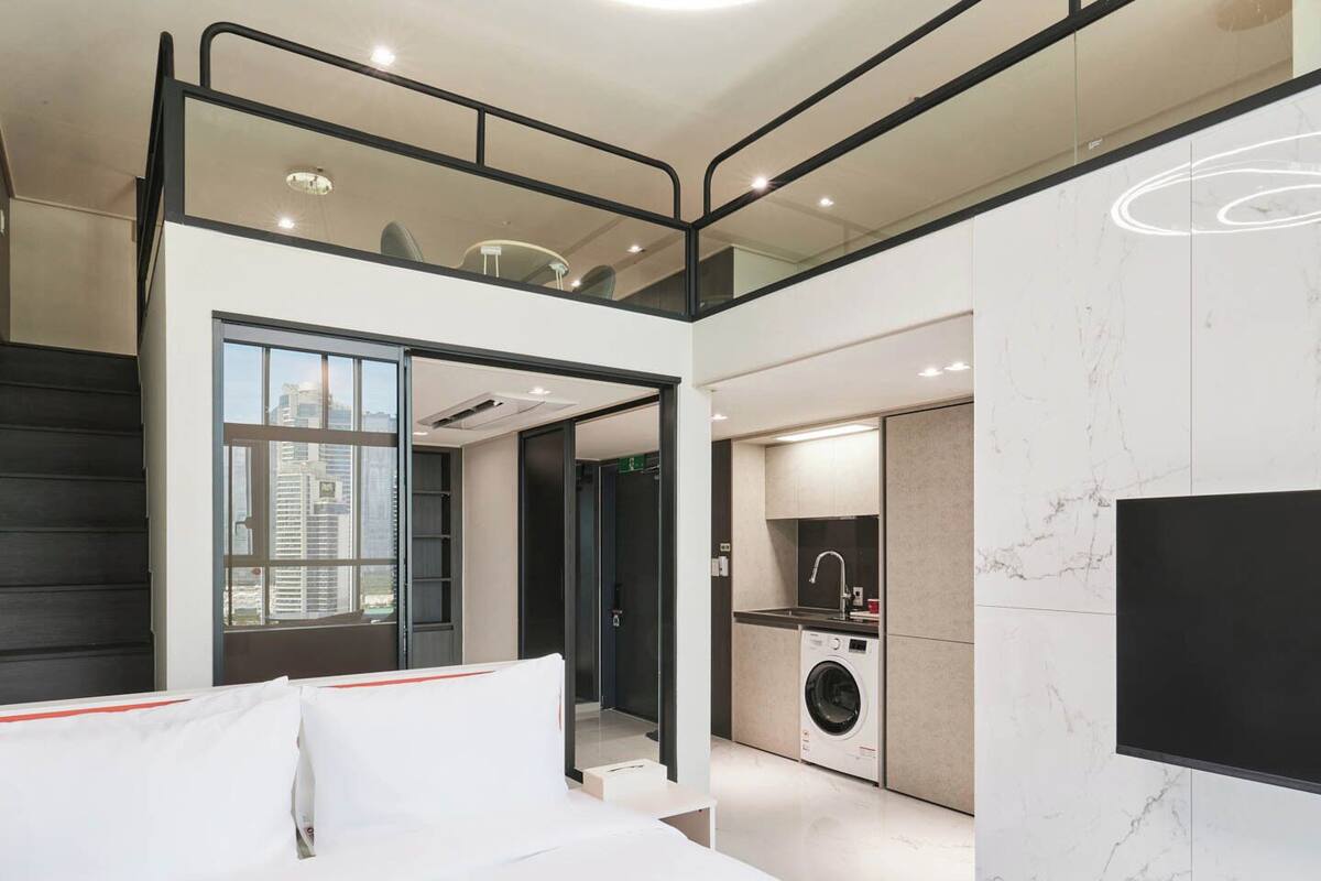 A modern hotel room with a bed, a built-in washer and dryer, a balcony, and a large window offering a city view.