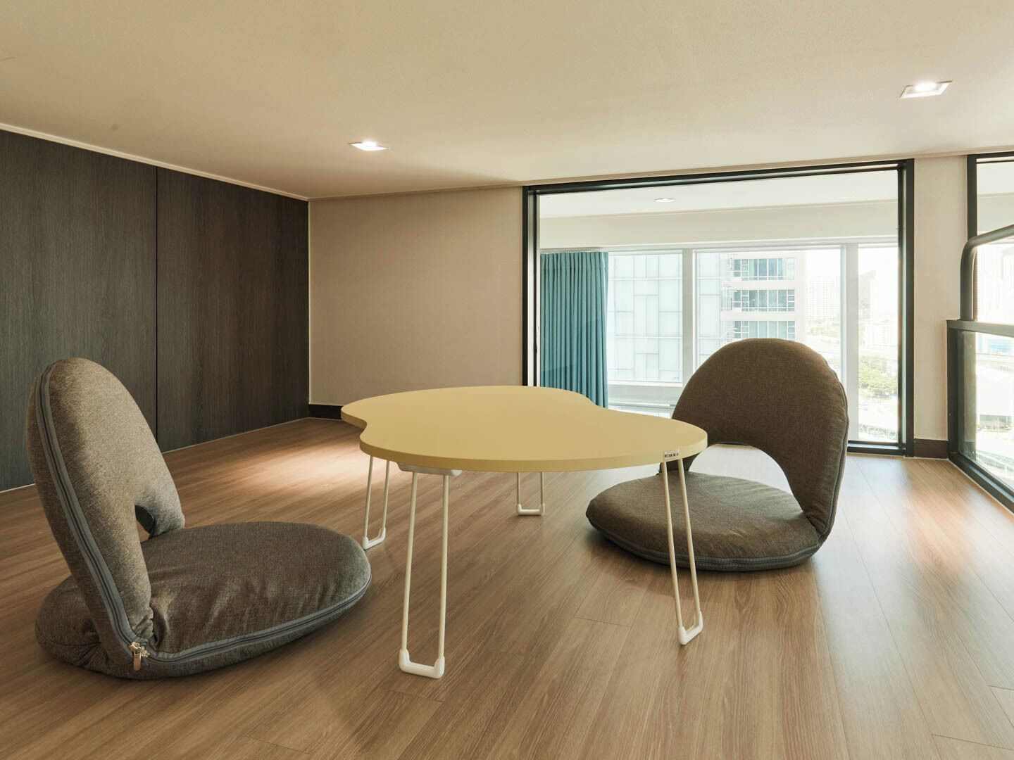 A modern room with a round table and two chairs, large windows with a view of buildings, and wooden flooring.