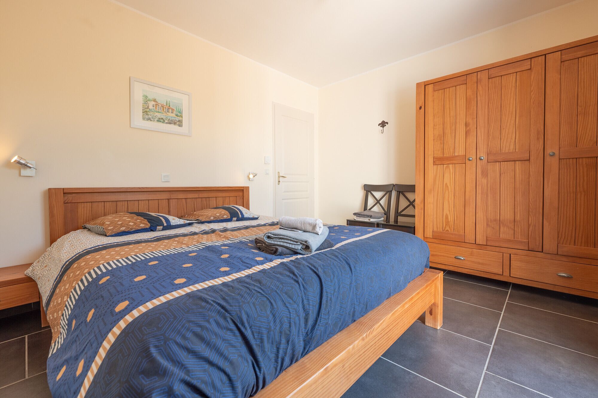 A bedroom with a large bed, a wooden wardrobe, and a small table with a chair.