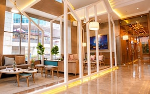 Un hall d’hôtel moderne doté d’un espace salon, d’un comptoir d’accueil et de grandes fenêtres offrant une vue sur le paysage urbain.