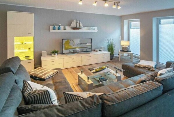 Uma sala de estar moderna com um sofá, uma mesa de centro, uma TV sobre um móvel branco e uma planta.