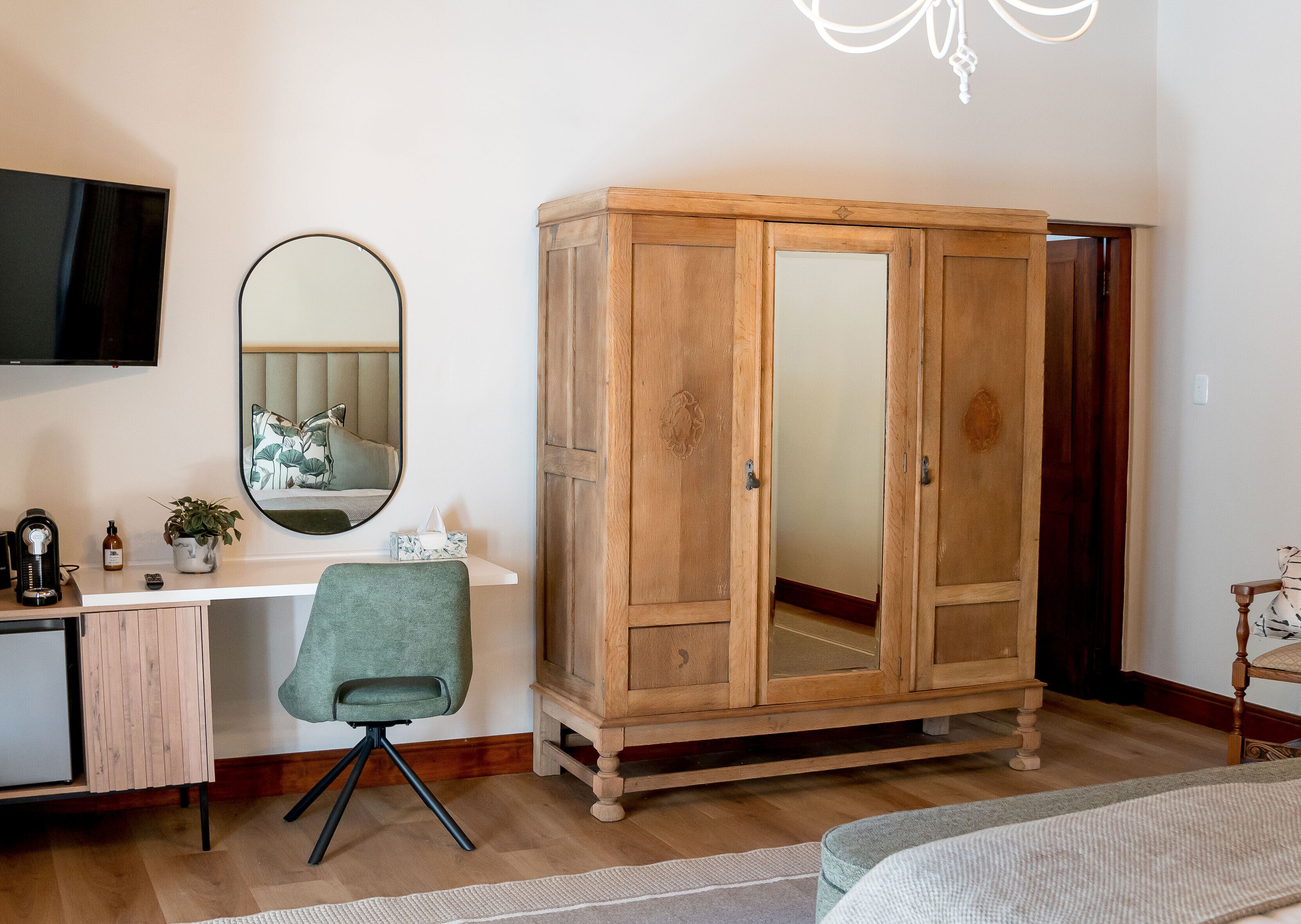 A bedroom with a wooden wardrobe, a desk with a chair, a television, and a mirror.