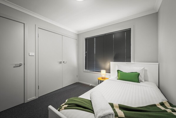 A bedroom with a bed, a nightstand, a window with blinds, and a door.