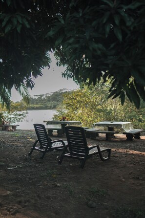 A serene outdoor seating area with a table and chairs under a tree, overlooking a calm body of water. - Dominical Long Term Rental (Bahía Ballena)