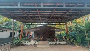 A covered outdoor area with a wooden structure, surrounded by plants and a small building in the background. - Dominical Long Term Rental (Bahía Ballena)