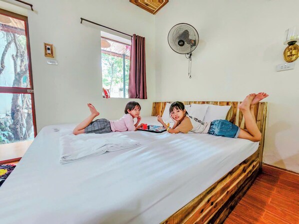A room with a bed, two children lying on it, a window with curtains, a wall-mounted fan, and a wooden headboard.