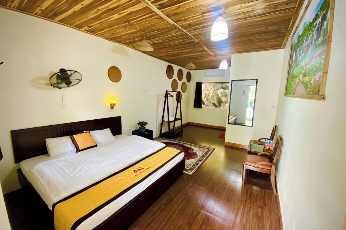 A hotel room with a wooden ceiling, a bed with a yellow bedspread, a small table with a chair, and a painting on the wall.
