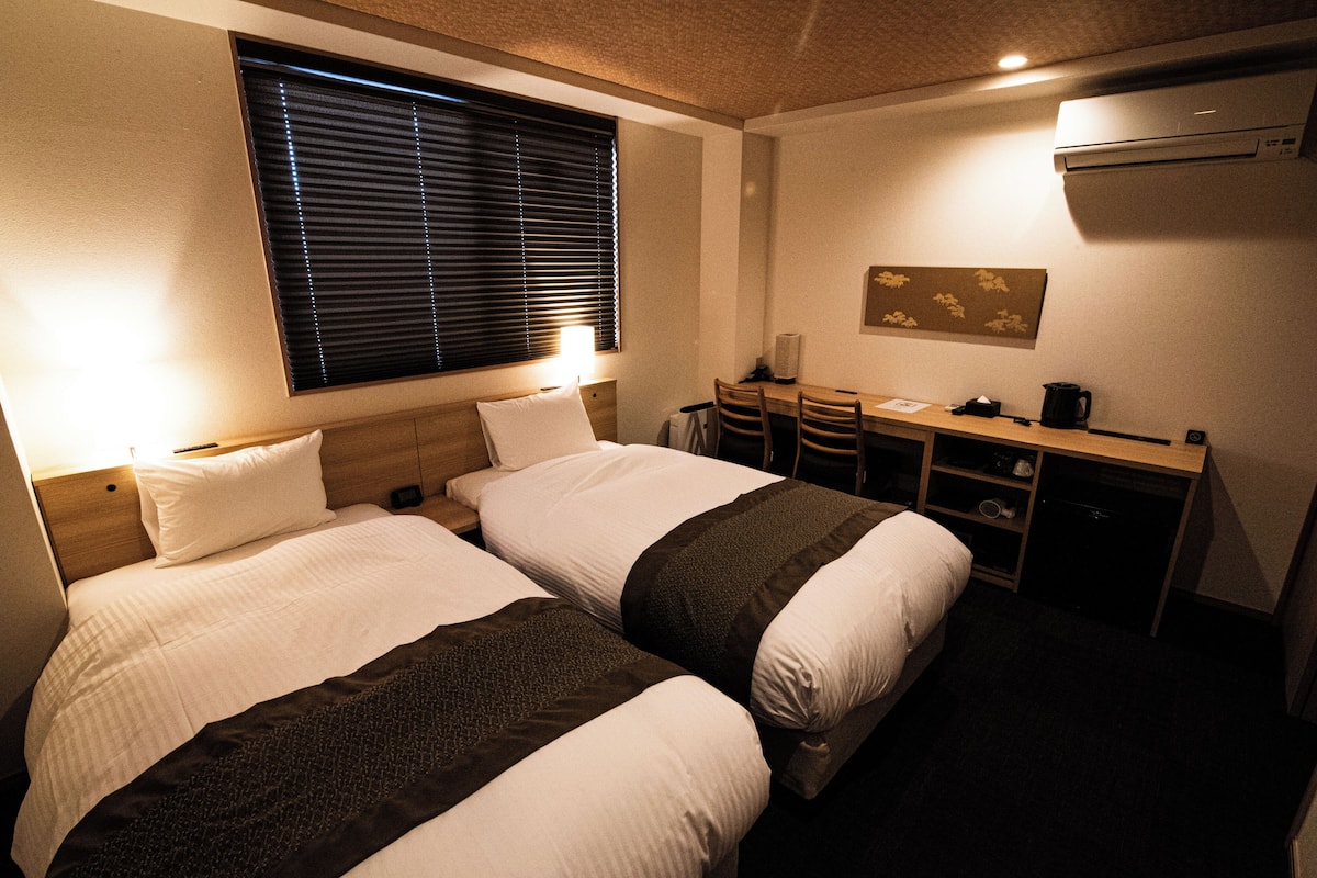 A hotel room with two beds, a desk, and a window with blinds.