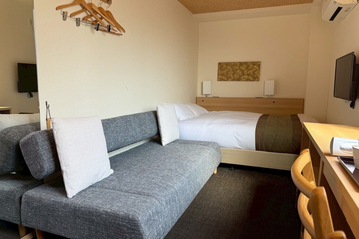 A hotel room with a grey sofa, a bed with white bedding, a wooden desk, and a television on the wall.