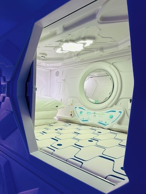 Una habitación con un ventanal circular, una cama con cabecera estampada y un panel de control con botones y pantalla.