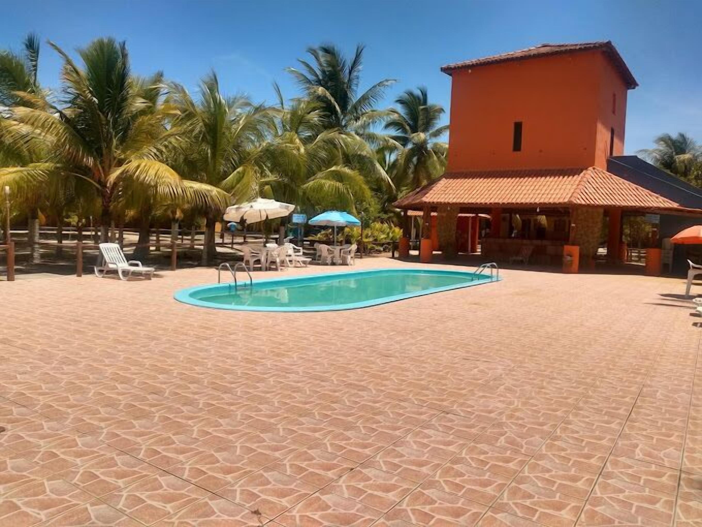 A swimming pool with lounge chairs and umbrellas, surrounded by palm trees and a building with a tiled roof.