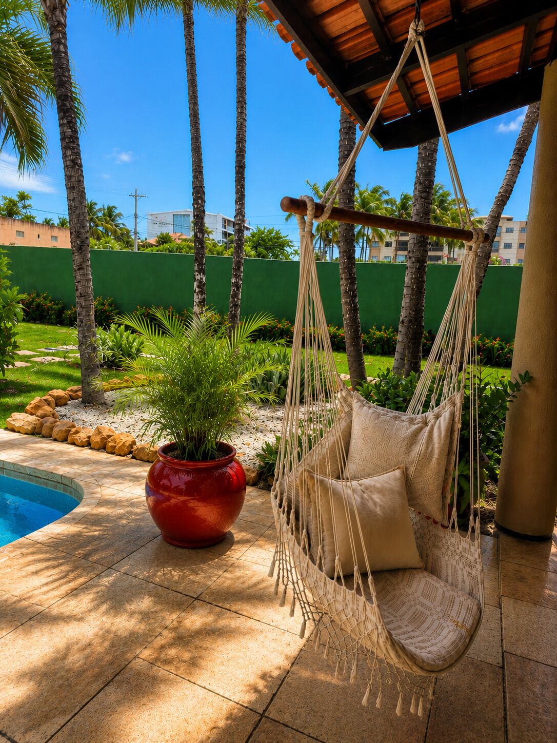 A hammock chair by a pool, surrounded by palm trees and a red pot.