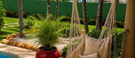 A hammock chair by a pool, surrounded by palm trees and a red pot.