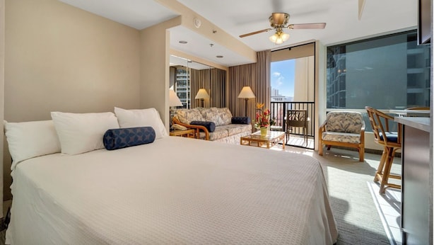 Habitación de hotel con una cama grande, un balcón con vistas, una zona de estar con mesa y un ventilador de techo.