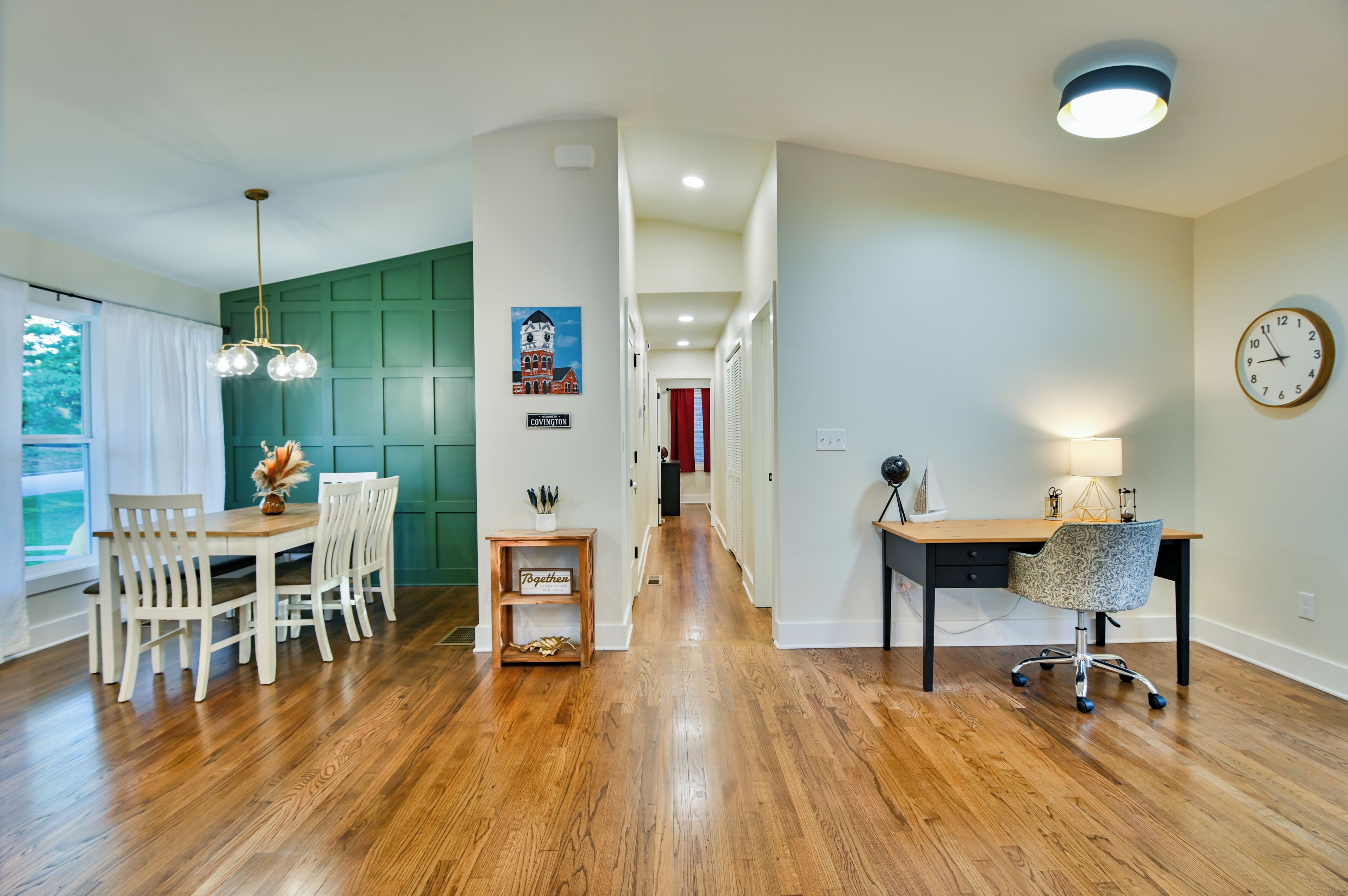 A modern kitchen with white cabinets, a dining area with a wooden table and chairs, and a hallway with a clock and a lamp.