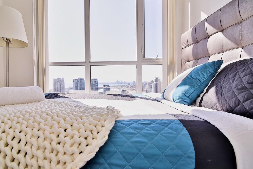 A modern bedroom with a large window, a city view, a tufted headboard, and a white knitted throw.