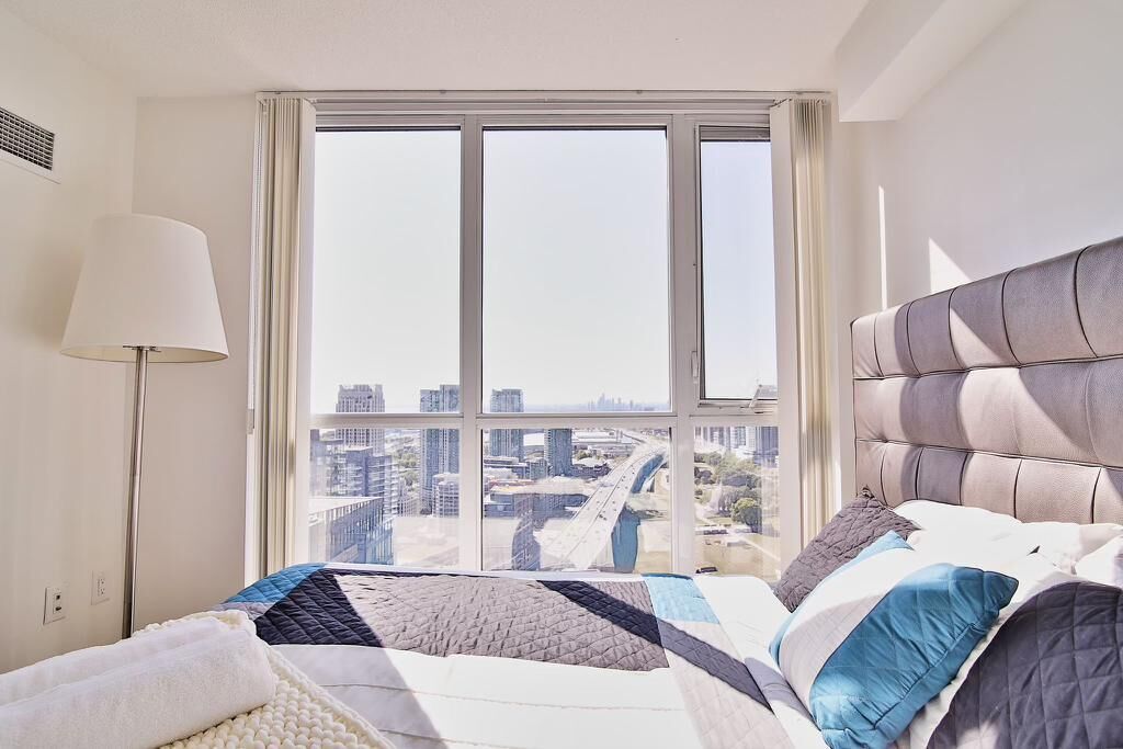 A bedroom with a large window, a bed with a grey headboard, and a floor lamp.
