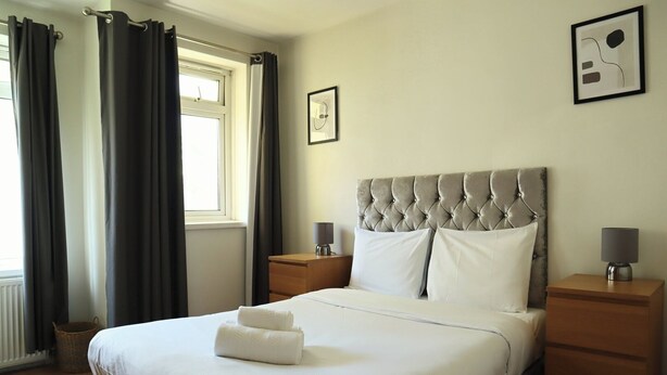 A neatly made bed with white linens and two pillows, a wooden nightstand with a lamp, a window with curtains, and a framed picture on the wall.