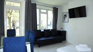 A modern hotel room with a black sofa, a blue armchair, a bed with white linens, and a large window with curtains.