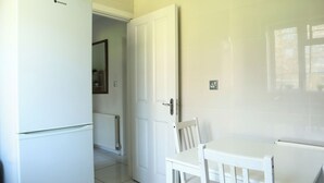 A kitchen with a white refrigerator, a white table, and a white chair. There is a door leading to another room.
