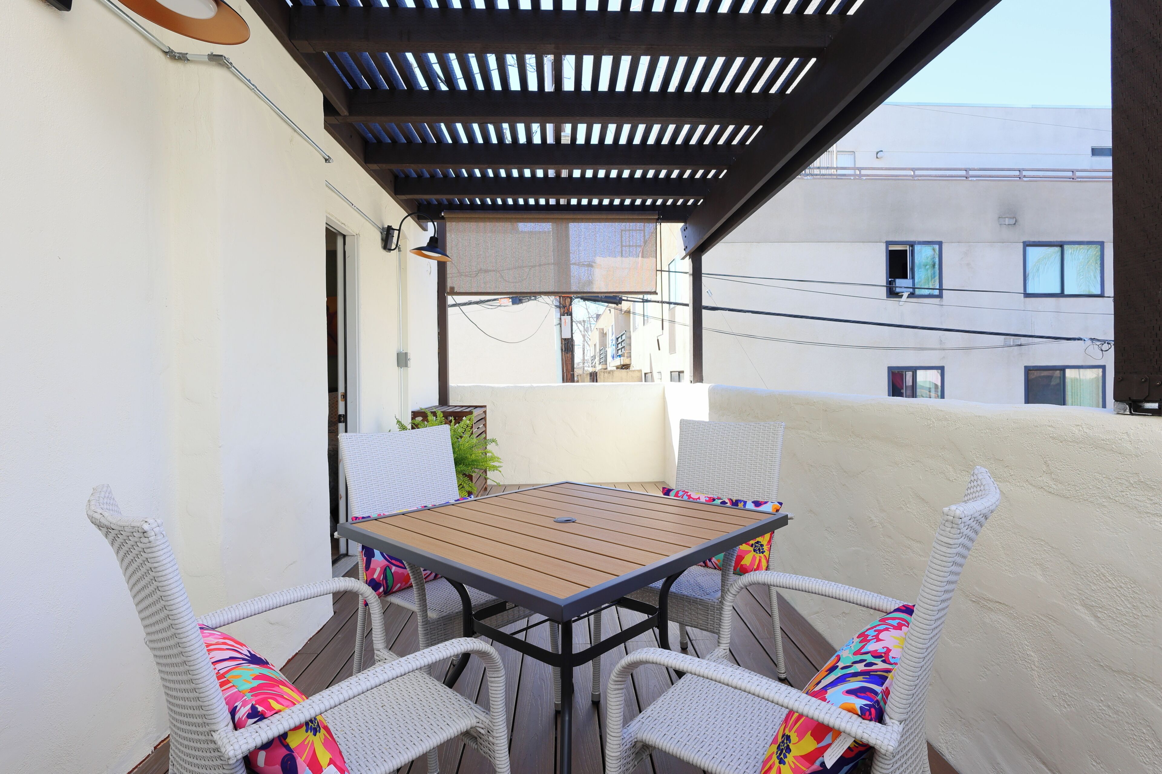 Un balcón con mesa de madera y sillas de mimbre, una pérgola arriba y vista de edificios al fondo.
