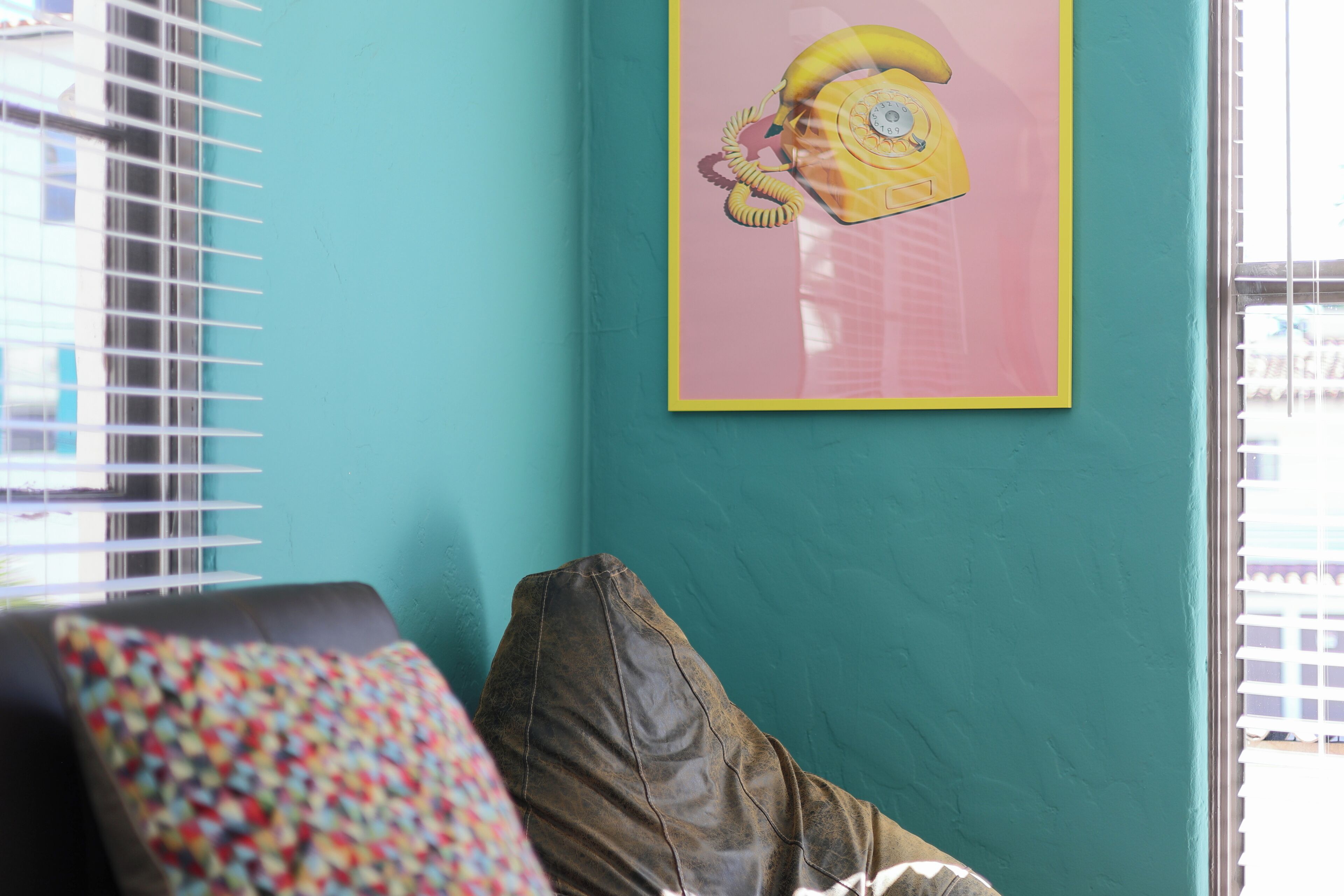 A teal-walled room with a framed yellow telephone art piece, a brown leather couch, and a colorful pillow.