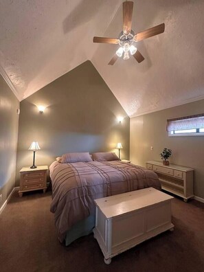 Un dormitorio con techo inclinado, una cama, dos mesitas de noche con lámparas, un tocador y un ventilador de techo.