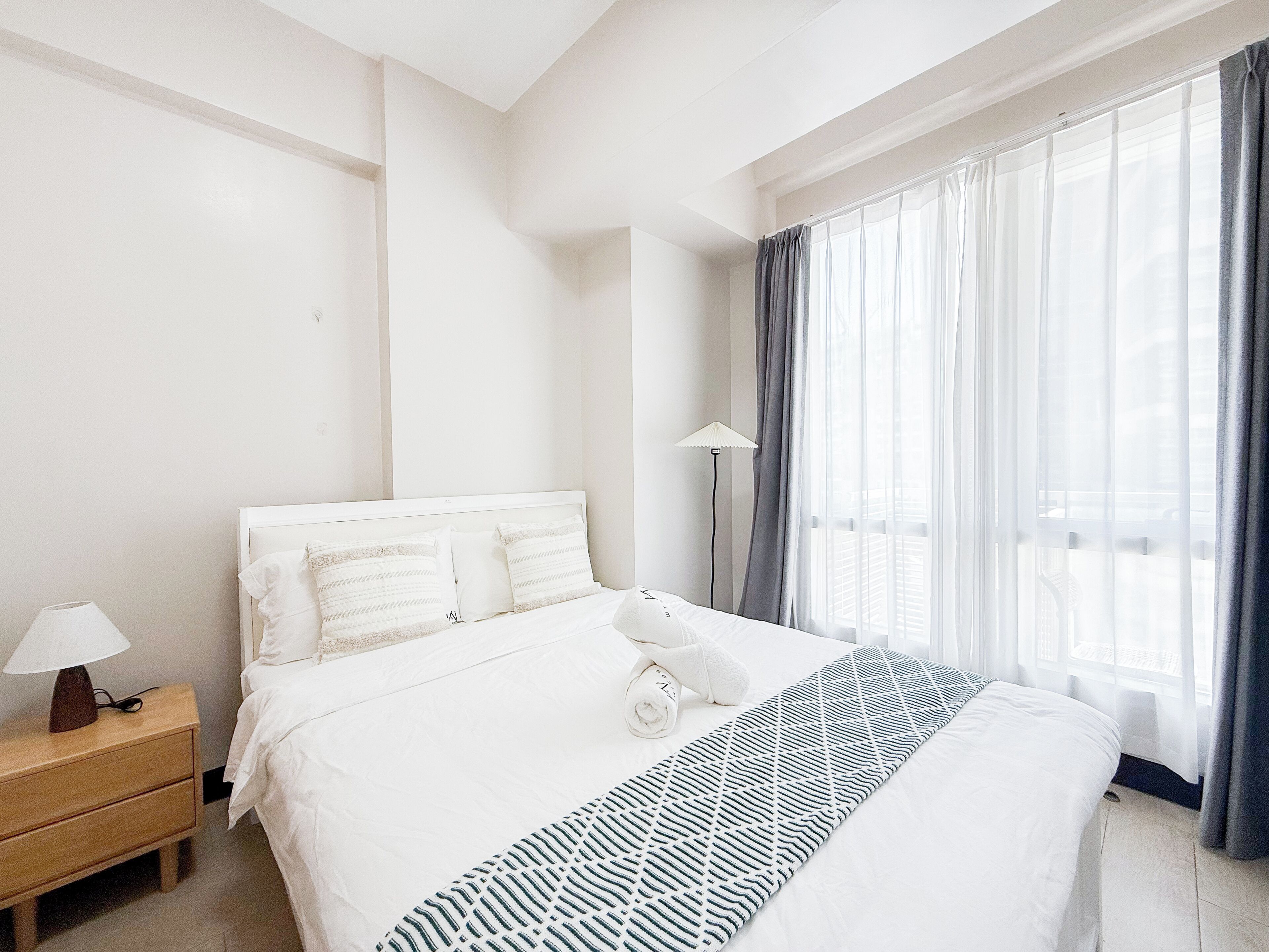 Habitació d'hotel amb un llit, una tauleta de nit, una làmpada i una finestra amb cortines.