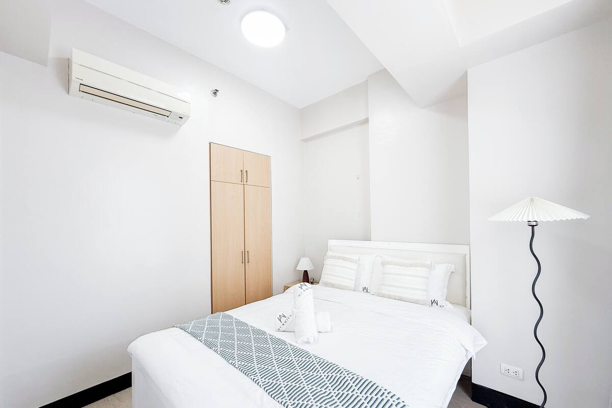 A hotel room with a bed, a wardrobe, an air conditioning unit, a lamp, and a power outlet.