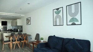 Een moderne woonkamer met een blauwe bank, een eethoek met houten stoelen en een keuken op de achtergrond.