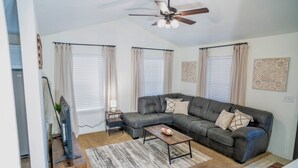 A living room with a sectional sofa, a coffee table, a TV on a wooden stand, a ceiling fan, and a large window with curtains.