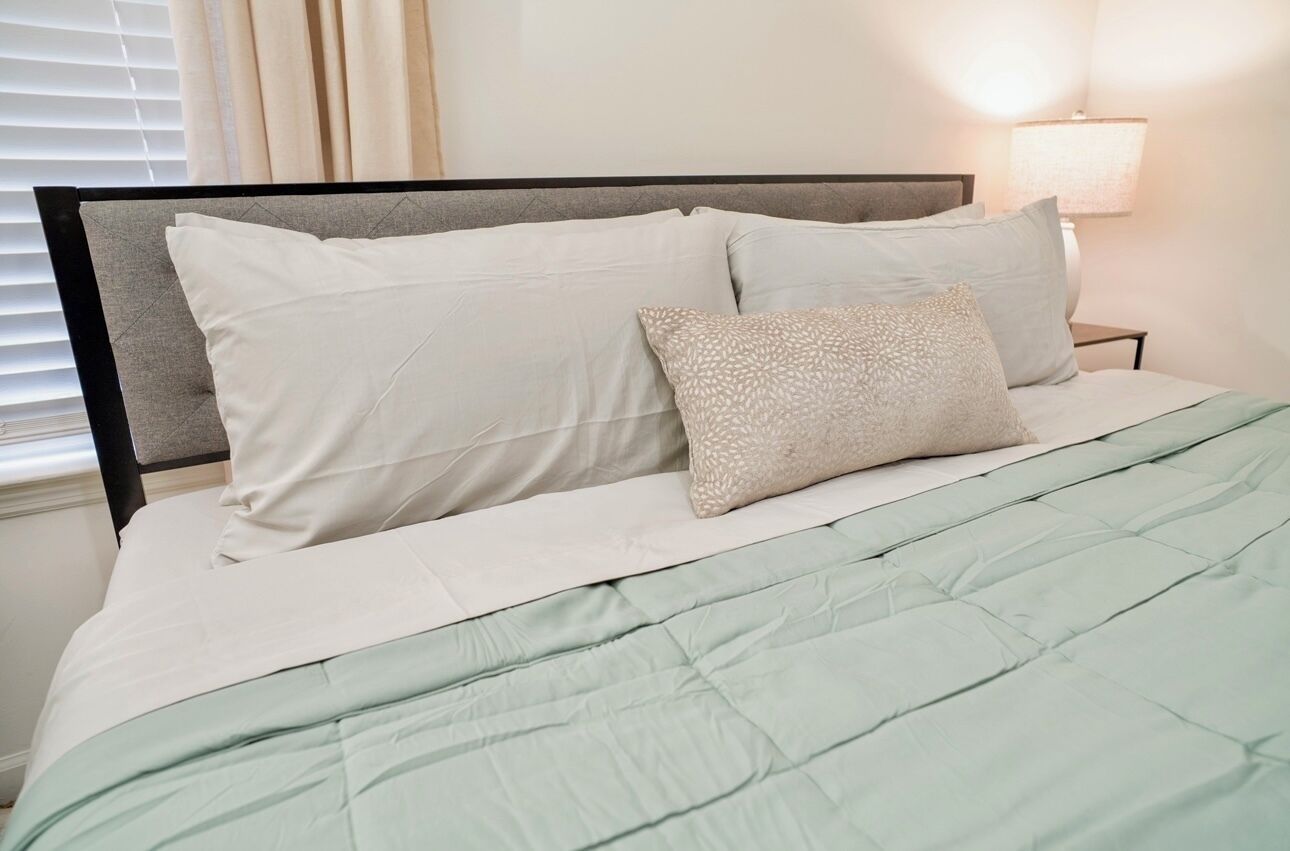 Un lit bien fait, recouvert d’une couette vert clair, avec une tête de lit grise et deux oreillers.